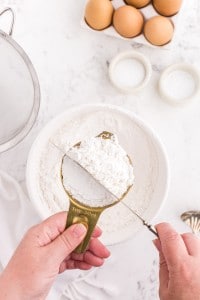 Sifted flour in a measuring cup being levelled by a knife, bowl of flour, sieve, tray of eggs, bowl of baking powder, bowl of salt, on a white marble countertop