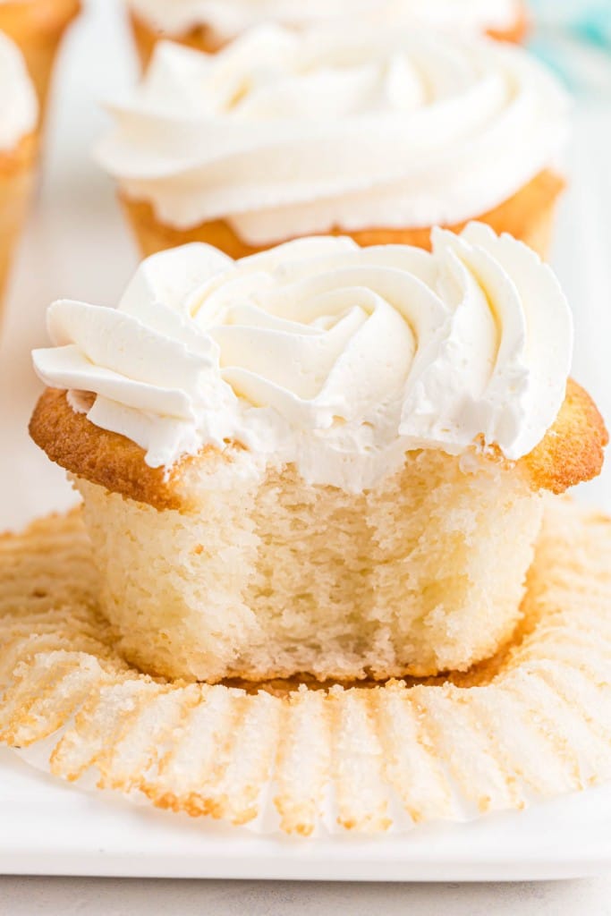 Close up of white cupcake with vanilla frosting and a bite taken out, additional cupcakes in the background, on a white plate