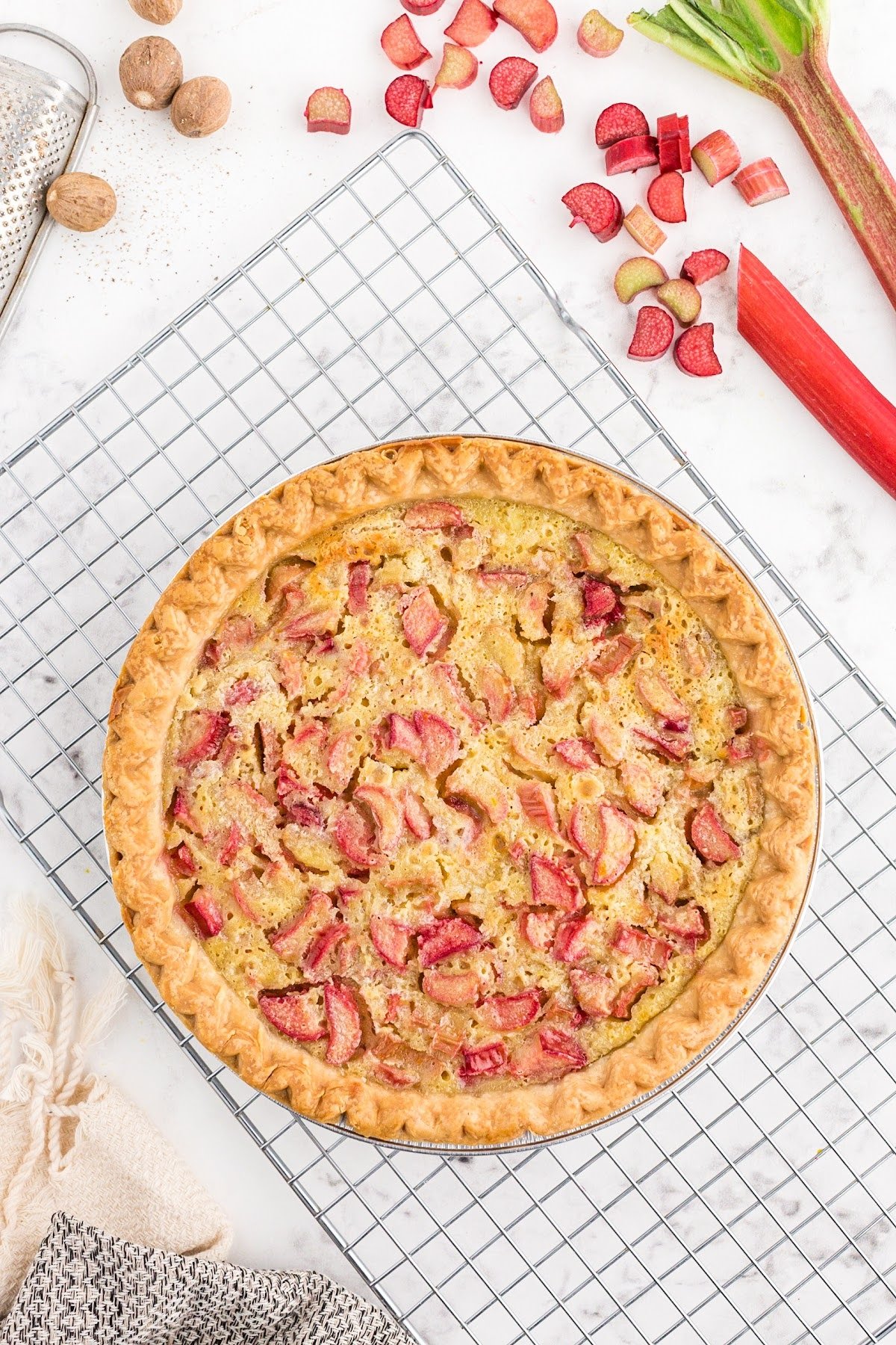 Baked rhubarb custard pie on metal cooling rack, linen, nutmeg, and rhubarb pieces scattered on counter.