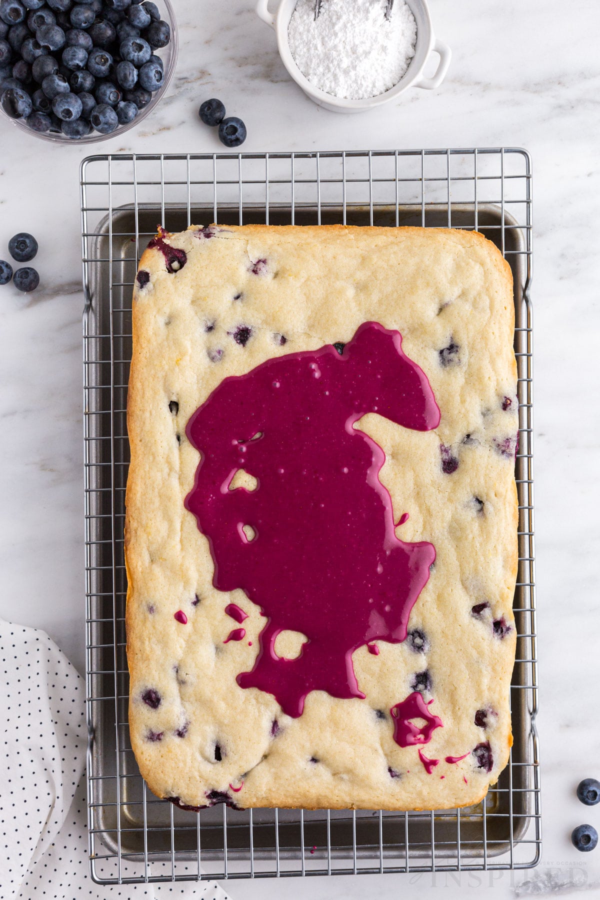 Baked blueberry lemon blondies on a wire cooling rack with metal tray underneath, blueberry glaze poured onto top of blondies, polka dot linen, on a white marble surface.