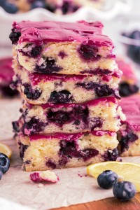 Stacked blueberry lemon blondies on parchment paper on a wooden kitchen board, white cake stand with blondies, bowl of fresh blueberries.