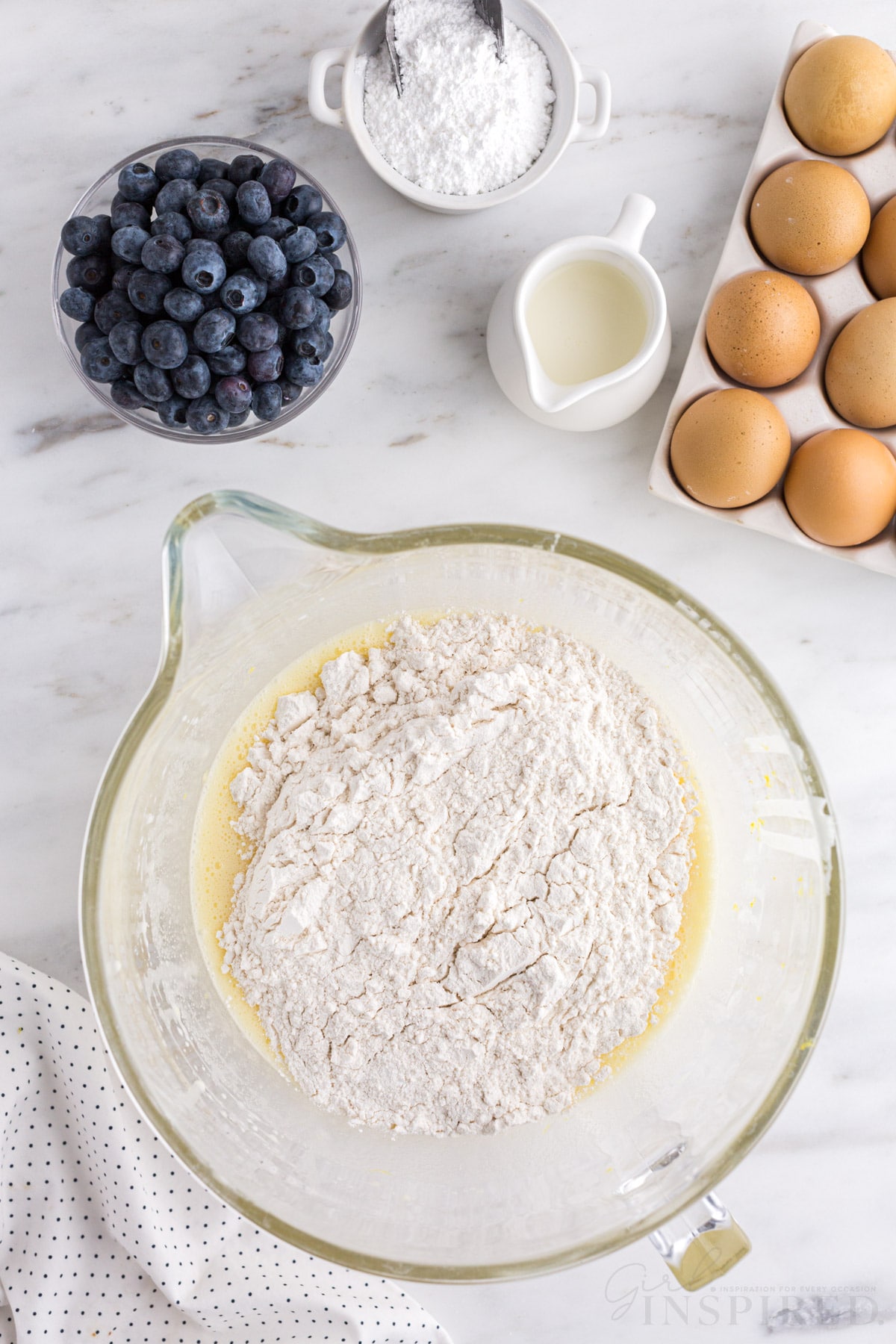 Glass mixing bowl with flour added to the mixed wet ingredients, bowl of fresh blueberries, tray of eggs, jug of milk, bowl of sugar, polka dot linen, on a white marble countertop.