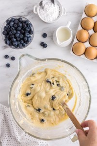 Fresh blueberries gently folded into the blondie batter in a glass mixing bowl, bowl of fresh blueberries, jug of heavy cream, tray of eggs, polka dot linen, on a white marble countertop.