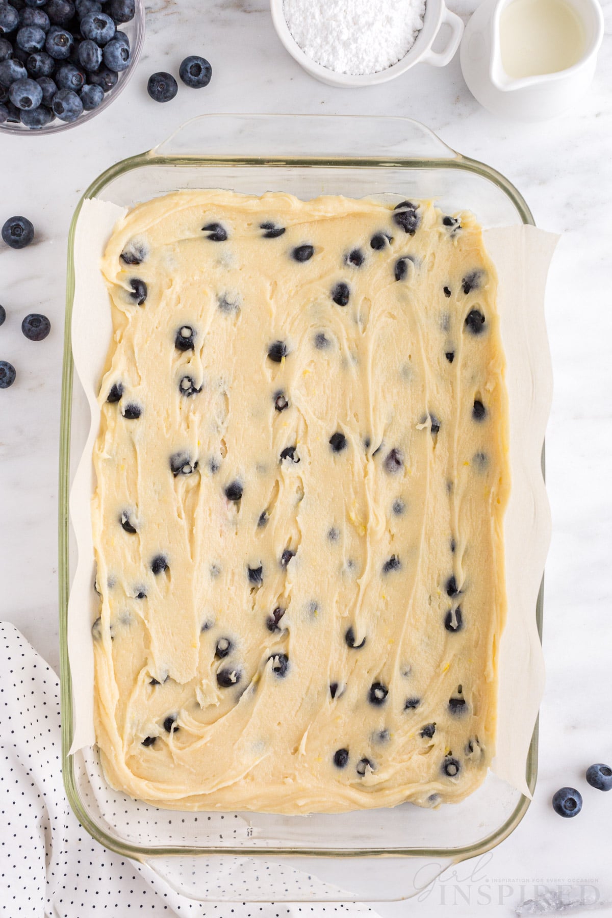 Glass baking dish lined with parchment paper and filled with blondie batter, fresh blueberries, polka dot linen, on a white marble countertop.