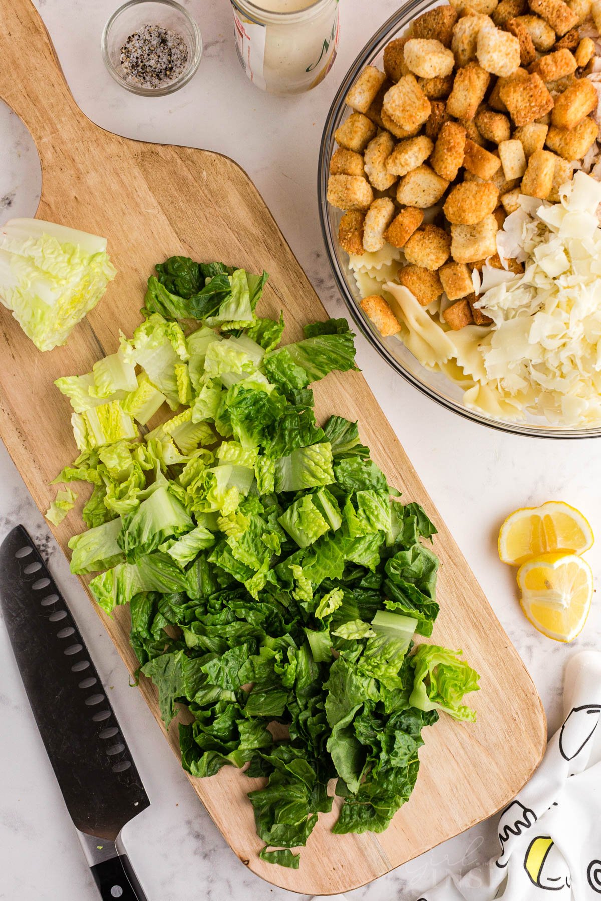 Chopped romaine lettuce on cutting board with a bowl of pasta, croutons, and cheese to the side.