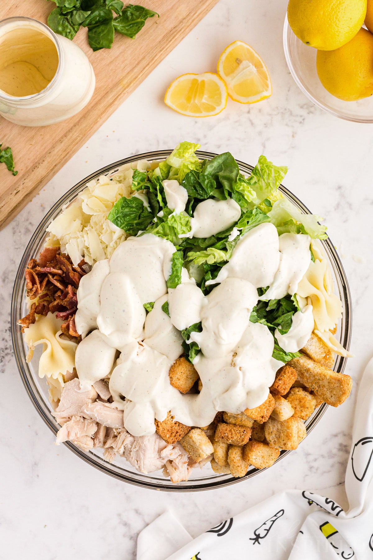 Caesar salad ingredients all added to a large glass bowl, placed on top of a white marble surface with excess salad ingredients scattered around.