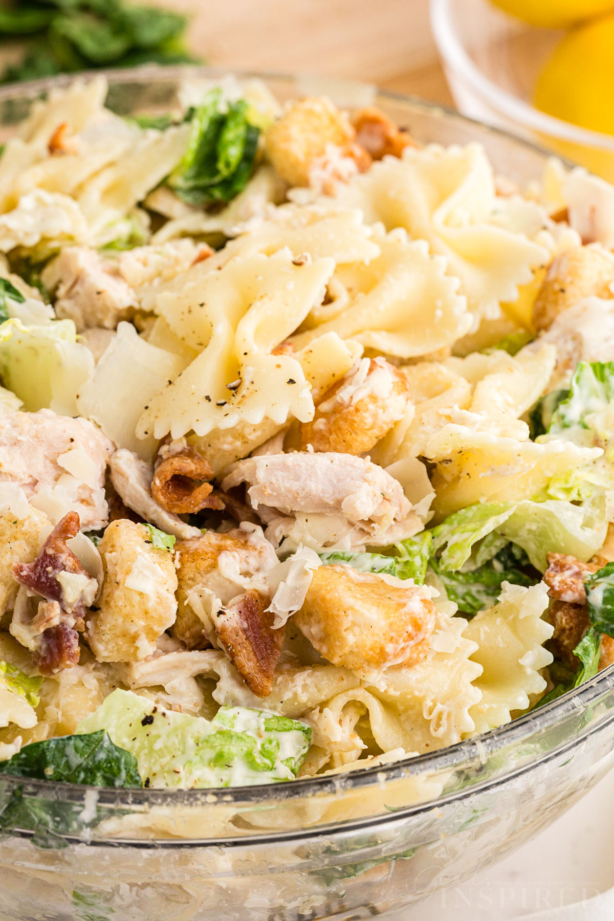 large glass serving bowl filled with chicken caesar pasta salad with wooden salad spoons and fresh lemons in the background, all placed on a white marble surface.