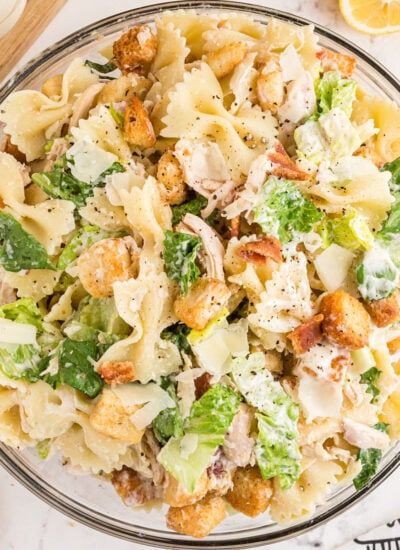 Large glass serving bowl filled with chicken caesar pasta salad with wooden salad spoons and fresh lemons in the background, all placed on a white marble surface.