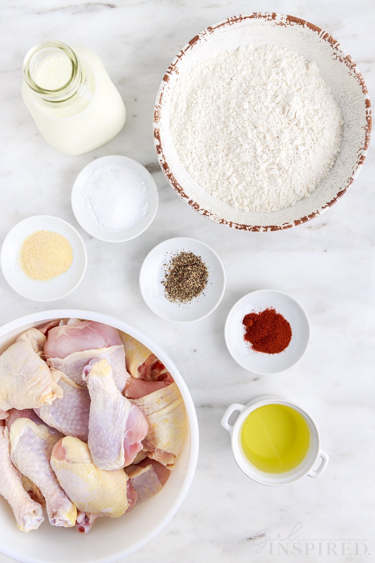 Bowl of chicken drumsticks and thighs, bowl of flour, jug of buttermilk, bowl of vegetable oil, bowls of seasoning, on a white marble countertop.