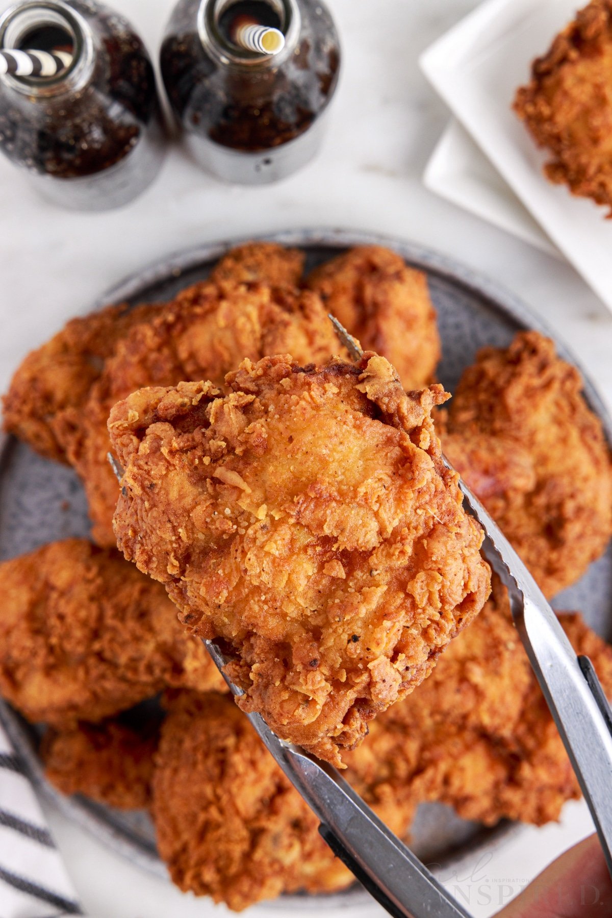 Fried chicken on a serving plate with metal tongs picking up a piece of fried chicken, navy striped linen, white serving plates with pieces of fried chicken, two bottles of soda with decorative straws on a white marble surface.