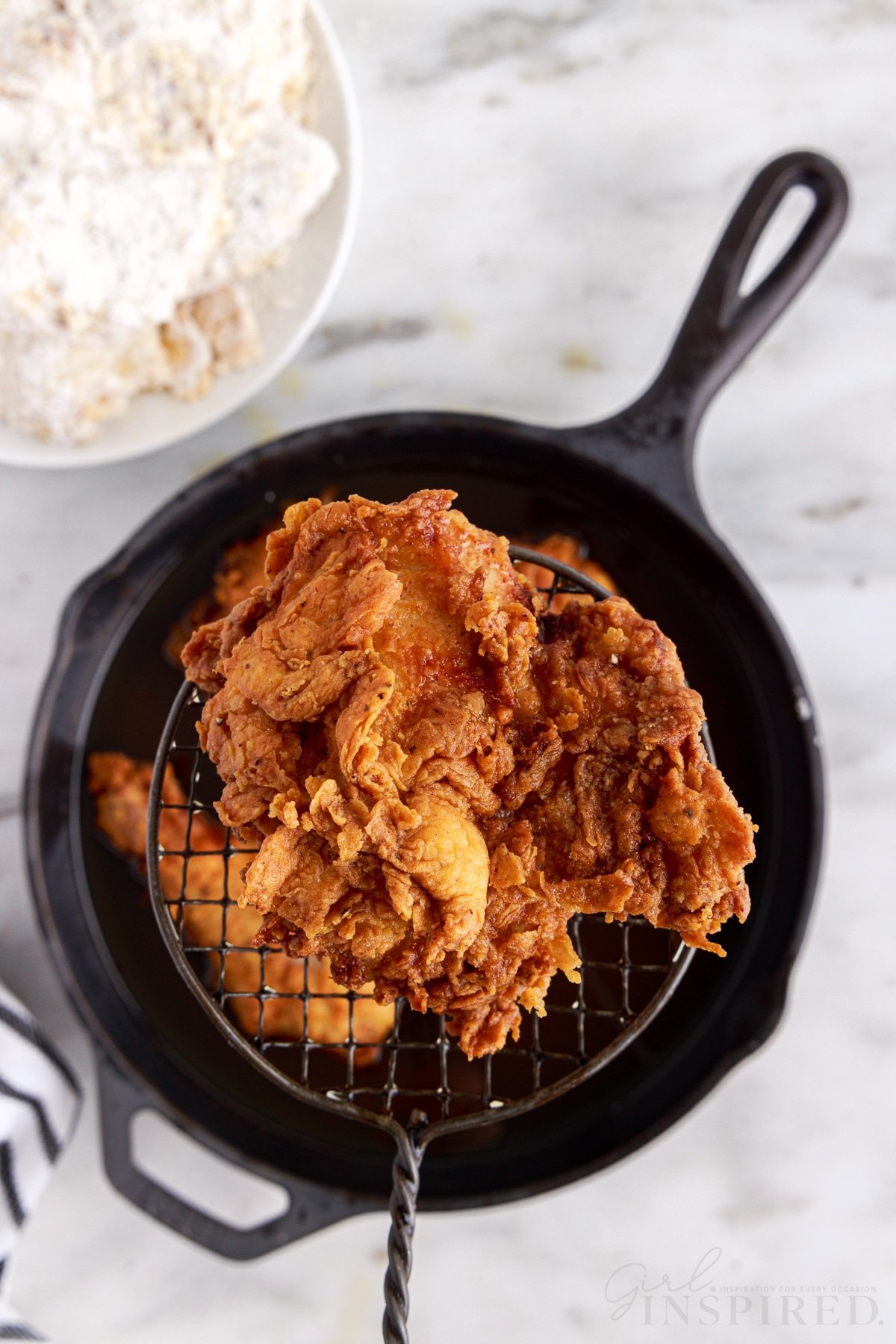 Fully deep fried chicken pieces lifted out of the Dutch oven with a slotted spoon, bowl of freshly battered chicken pieces, navy striped linen on a white marble surface.