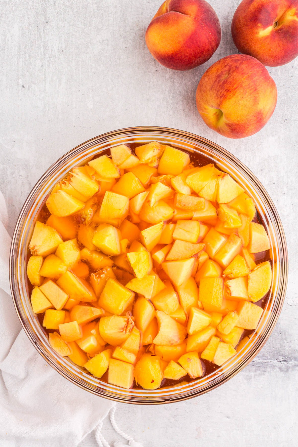Bowl of dissolved and set Jell-O with pieces of peaches, whole peaches, white linen, on a marble countertop.