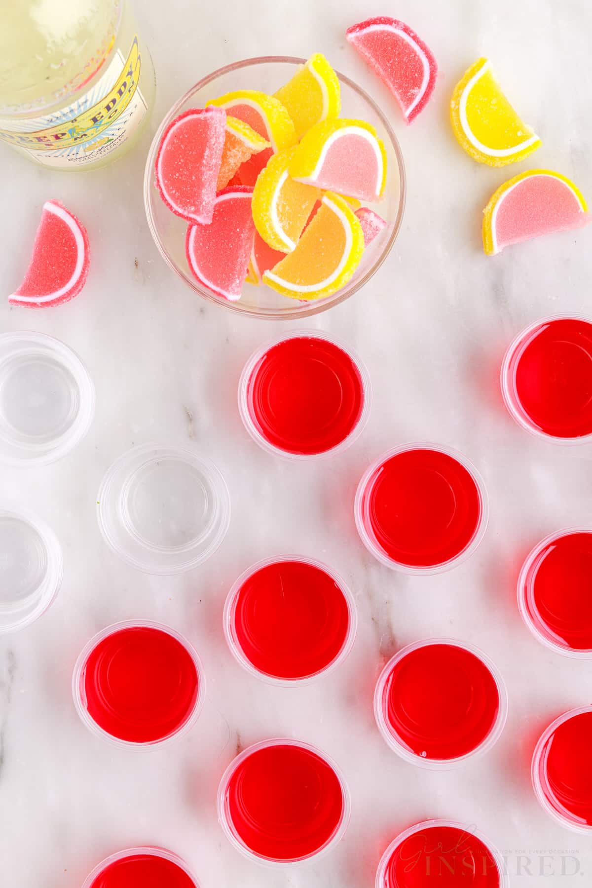 Pink lemonade jello shots in cups next to candies and other cups.