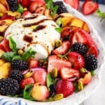 Ball of burrata cheese with sliced peaches and berries arranged around it on a serving platter. Peach burrata salad is drizzled with olive oil and balsamic glaze.