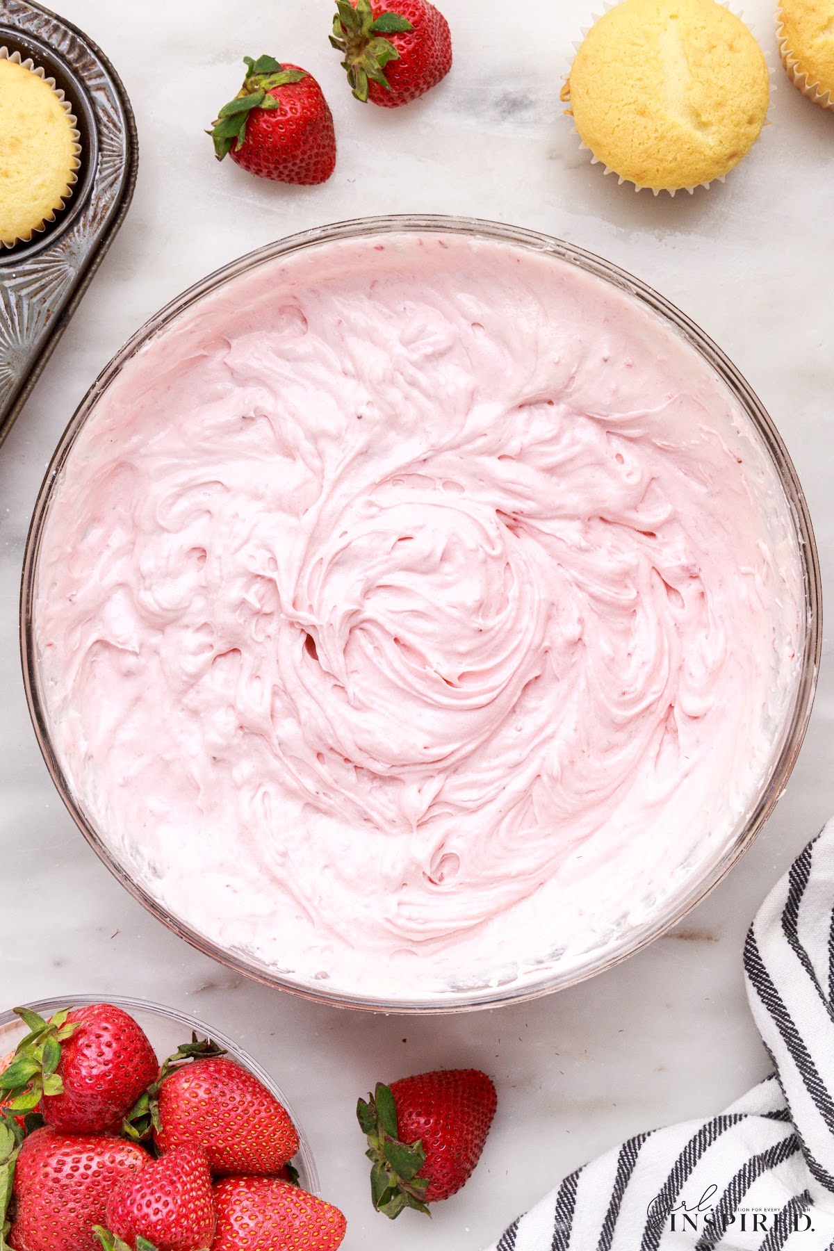 Large glass bowl of strawberry cream cheese frosting with cupcakes and whole strawberries scattered around.