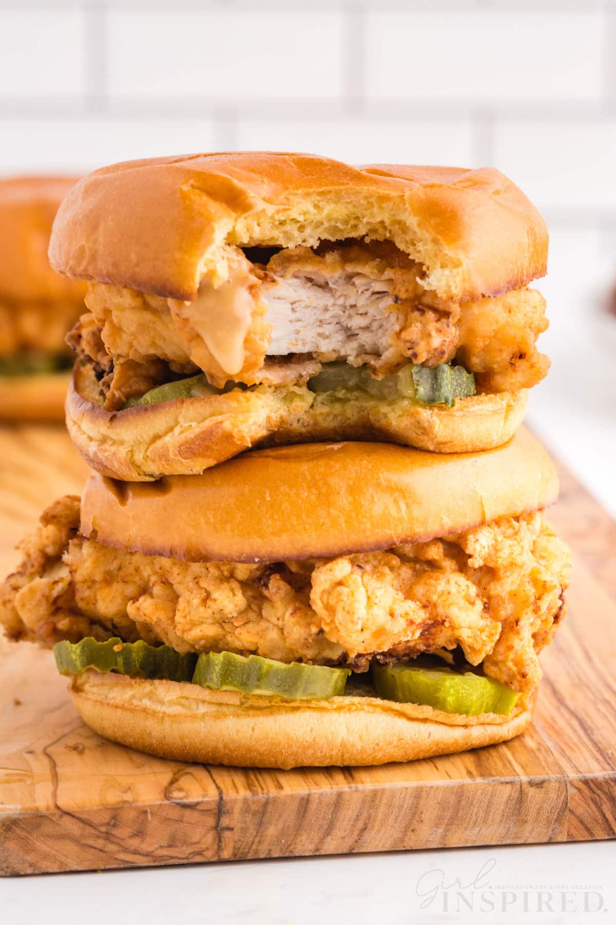Two Fried Chicken Sandwiches stacked on wooden kitchen board on marble countertop, white subway tiles in the background.