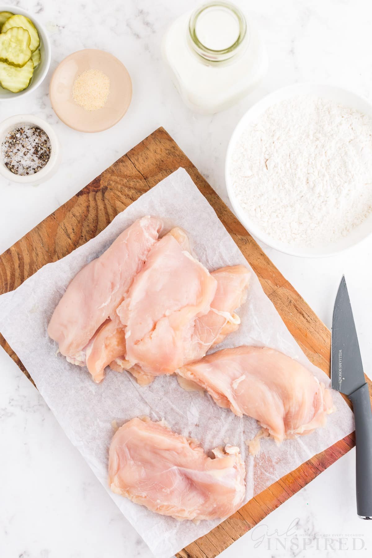 Several chicken breasts on wooden kitchen board lined with parchment paper, knife, bowl of flour, bowl of oil, bowl of sliced dill pickles, bowl of seasoning, bottle of buttermilk, on a white marble surface.