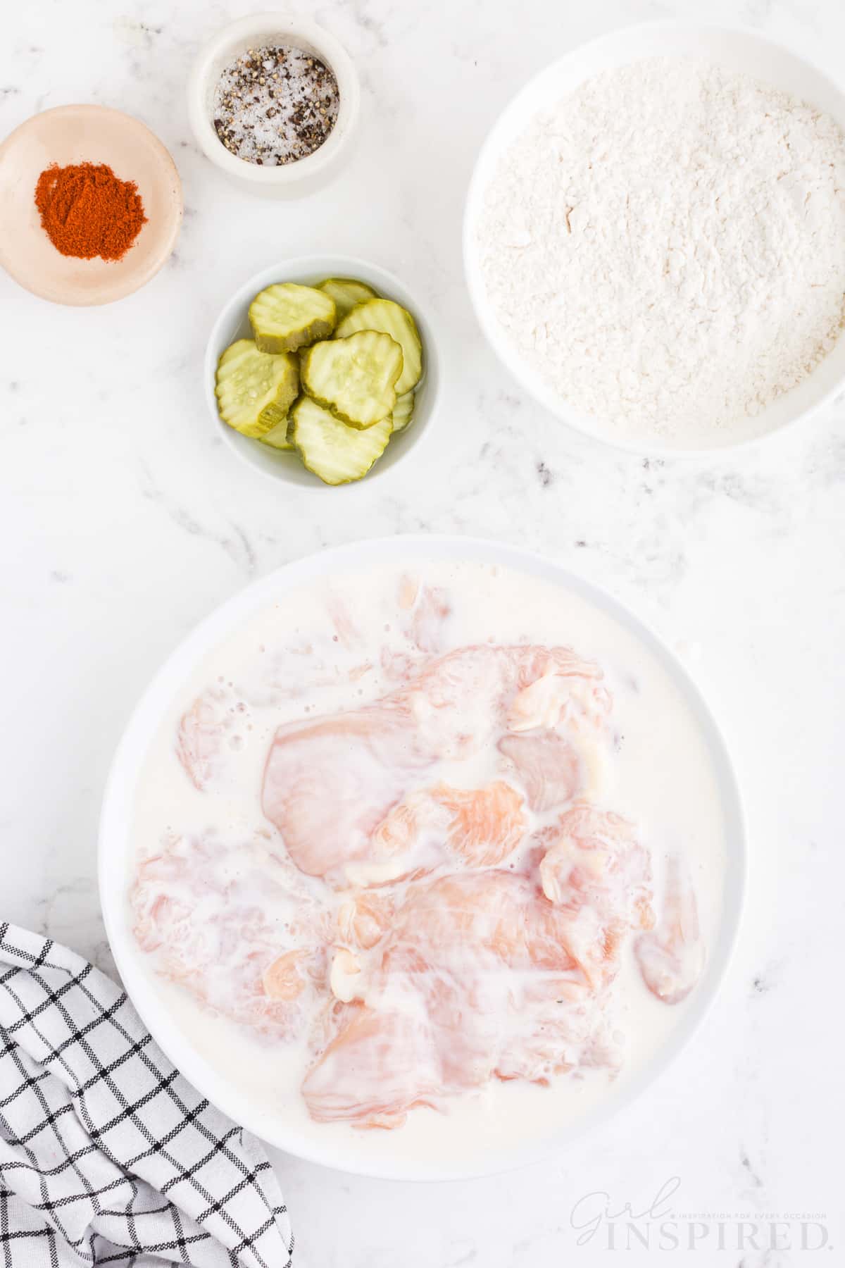 Bowl with buttermilk and raw chicken pieces, bowl of hot sauce, sliced dill pickles, flour, and seasoning, blue and white checked linen, on top of a white marble surface.