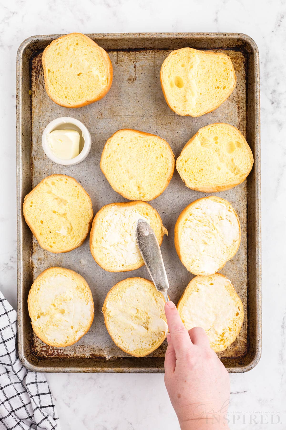 Spreading butter on open burger buns with knife on baking sheet, black and white linen, on a white marble surface.