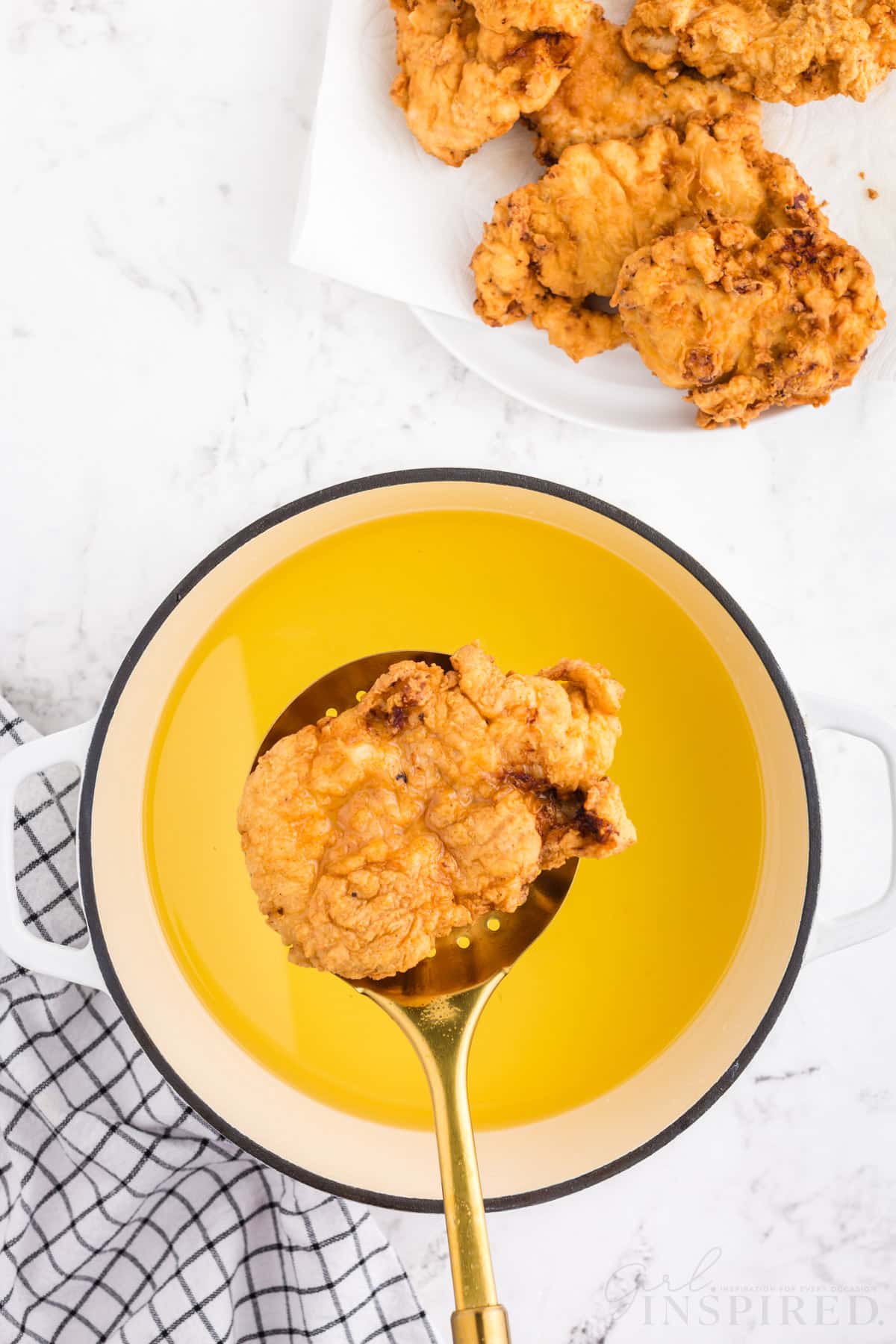 Fried chicken breast on slotted spoon over Dutch oven filled with vegetable oil, blue and black checked linen, serving plate of crispy fried chicken pieces, on a white marble surface.
