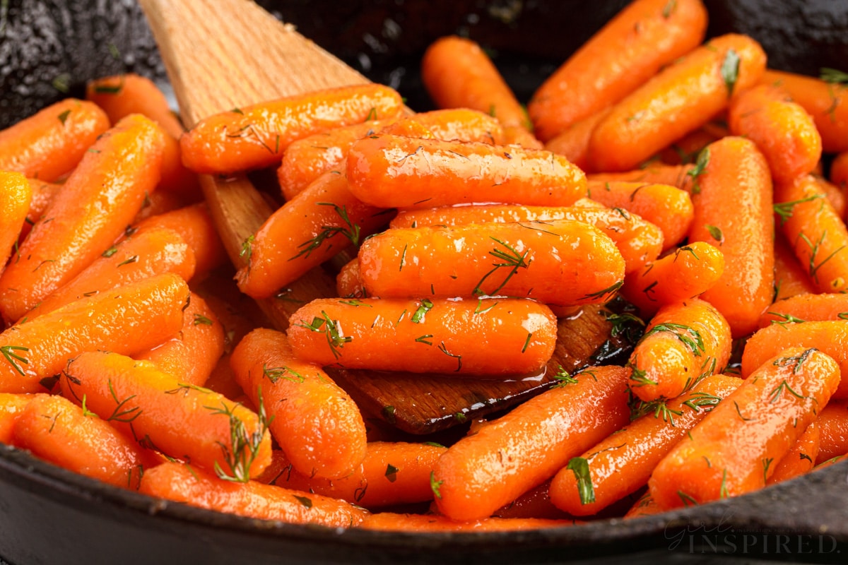 close up of brown sugar honey glazed carrots with wooded spatula raising a few off the skillet
