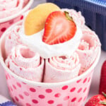 Pink polka dot cup with strawberry rolled ice cream, whipped cream, a strawberry, and a cookie on top with blue berry basket in background.