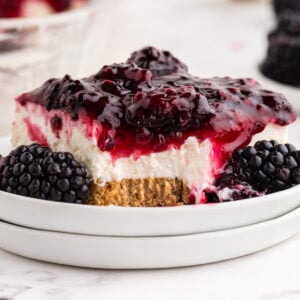 Slice of no bake blackberry cheesecake on staked white serving plates with fresh blackberries, forkful of dessert removed from the slice, no bake blackberry cheesecake in the background, striped linen, on a marble surface.