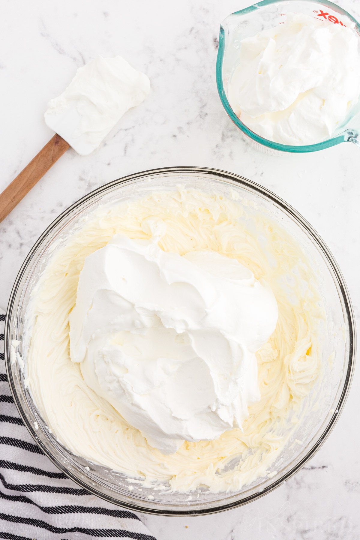 Whipped cream added to bowl of cream cheese mixture, striped linen, bowl of whipped cream, rubber spatula, on a white marble surface.