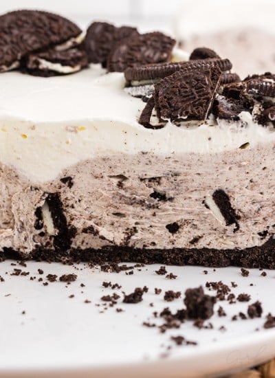 No bake Oreo cheesecake with slices removed on a white plate, on top of a woven mat, single slice of no bake Oreo cheesecake on staked white side plates in the background