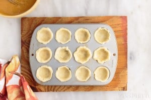 a 12 cavity mini muffin tin with each hole lined with pumpkin pie bites dough on top of a wooden cutting board