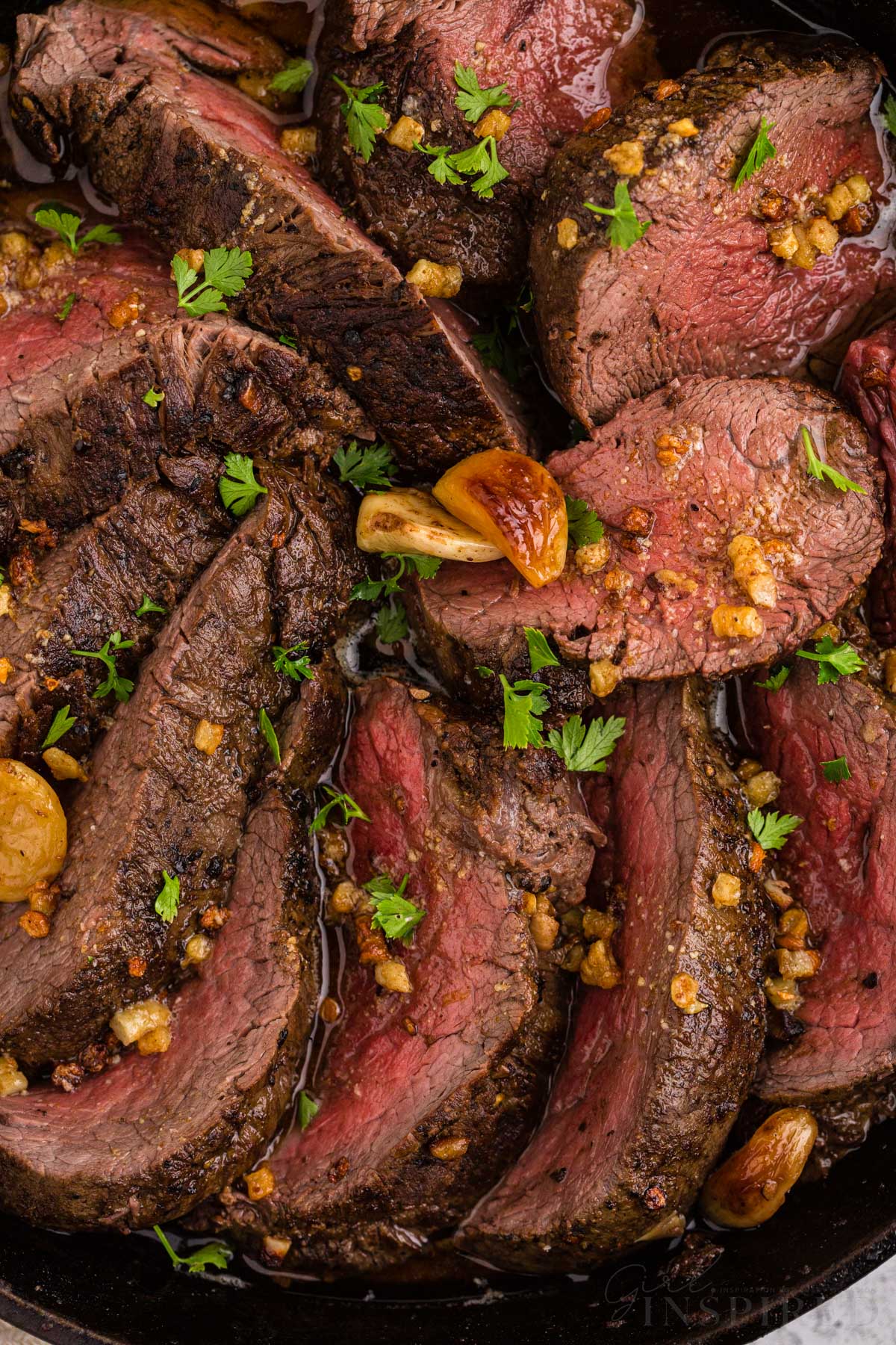 Close up of sliced roasted beef tenderloin in skillet with herb and roasted garlic garnish.