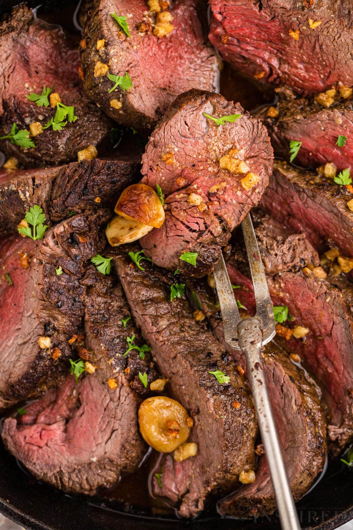 Close up of sliced roasted beef tenderloin in skillet with herb and roasted garlic garnish.