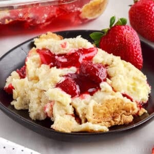 A serving of strawberry cheesecake dump cake on a black plate atop a white marble surface with baking dish and cloth in the background.