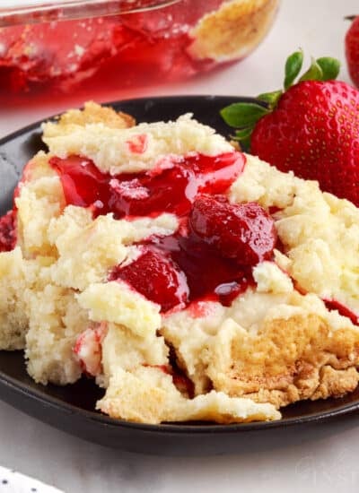 A serving of strawberry cheesecake dump cake on a black plate atop a white marble surface with baking dish and cloth in the background.