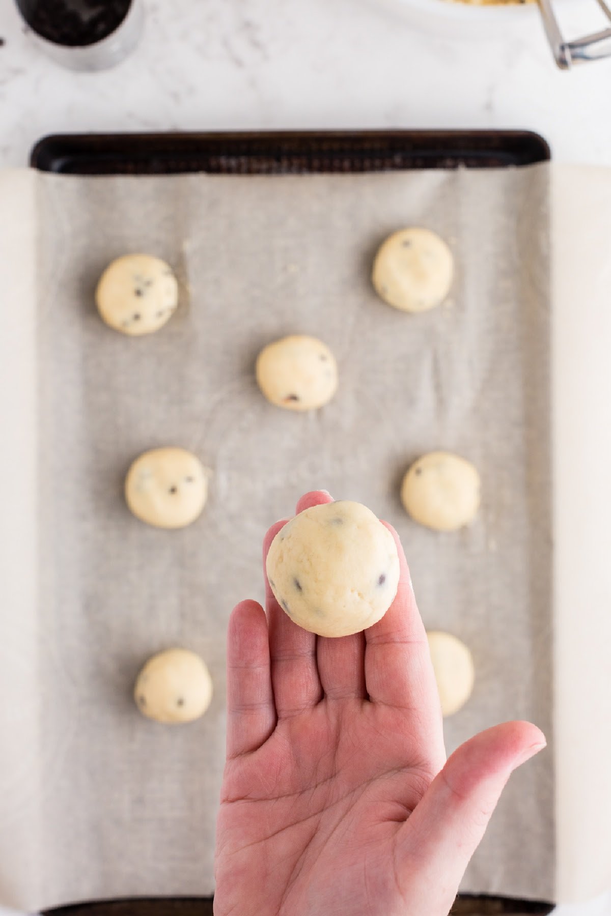 Smooth ball of chocolate chip cheesecake cookie dough held in the palm of hand over tray of cookie dough balls.