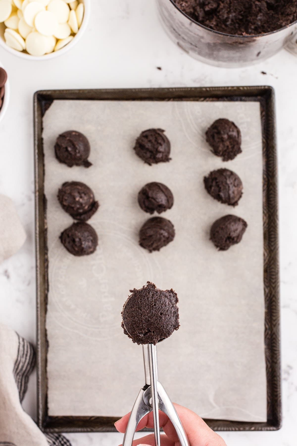 Cookie scoop measuring Oreo ball mixture, Oreo balls added to a parchment-lined baking sheet, bowl of dark chocolate melting wafers, bowl of white chocolate melting wafers, beige linen, on a white marble surface