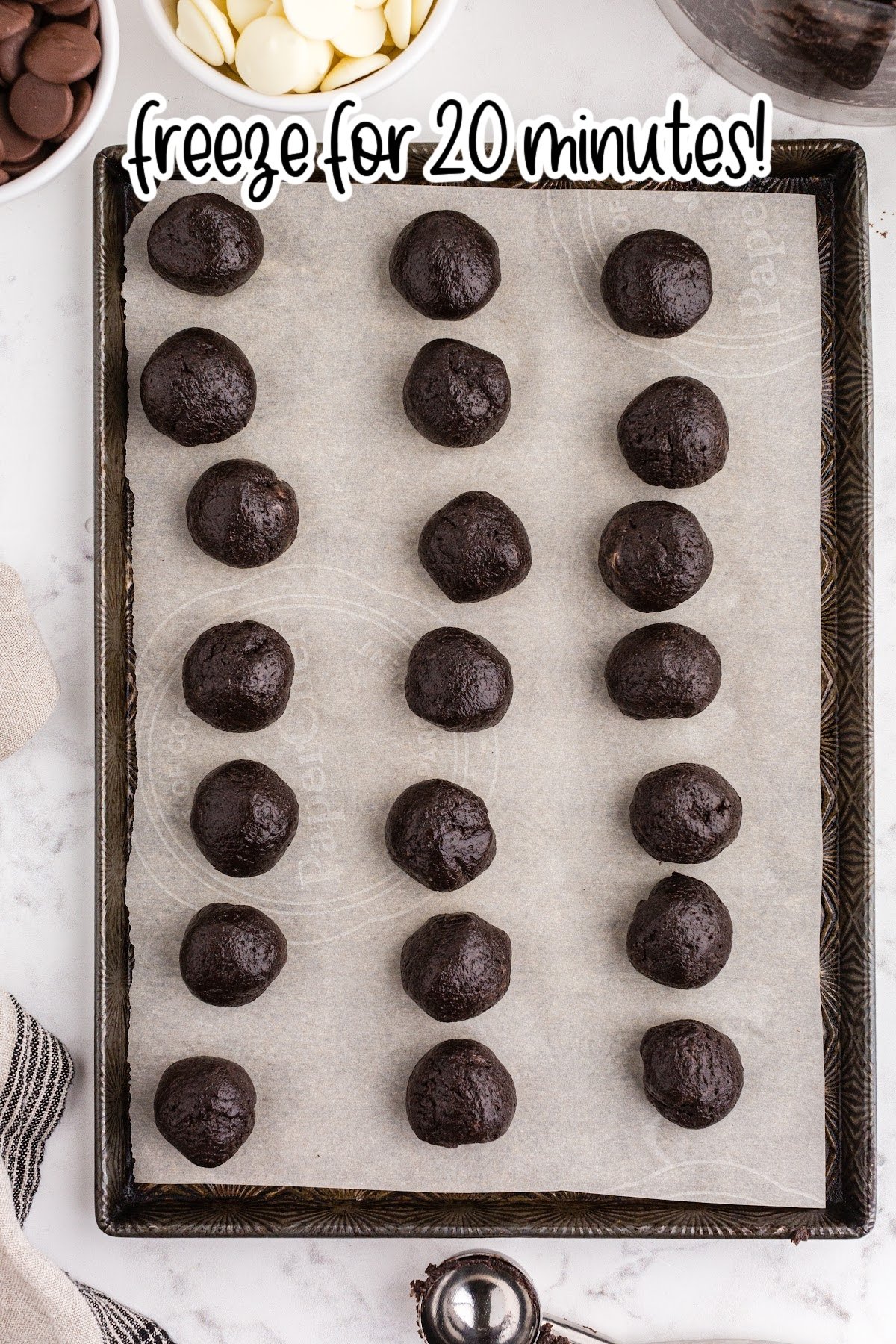 Parchment paper-lined baking sheet with frozen Oreo balls, metal cookie scoop, bowl of dark chocolate melting wafers, bowl of white chocolate melting wafers, beige linen, on a white marble countertop