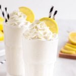 Two glasses of whipped lemonade topped with big swirl of whipped cream, a lemon wheel, and striped straws.