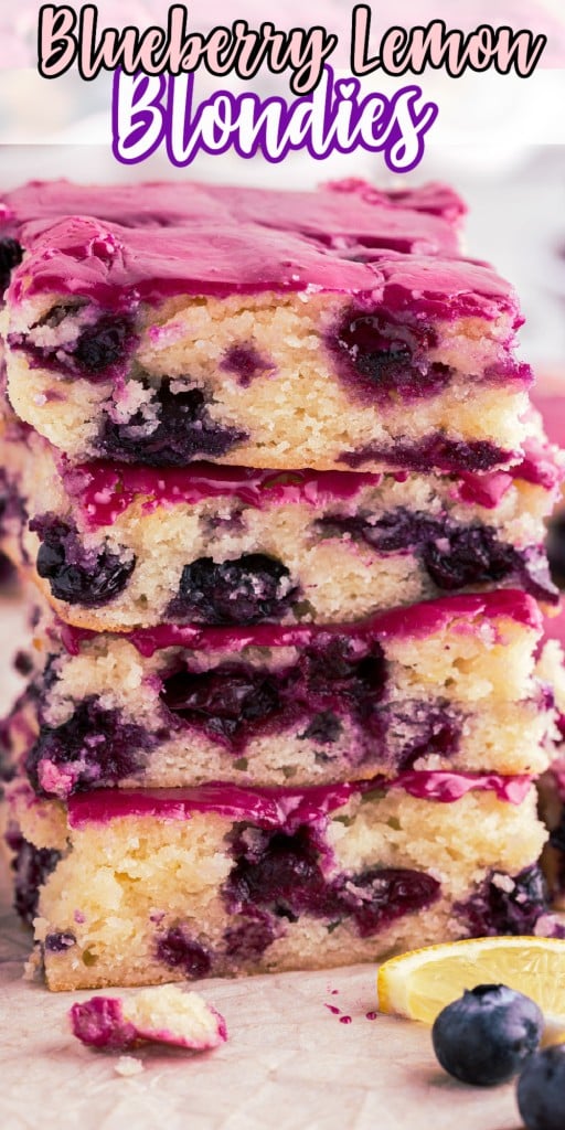 Stacked blueberry lemon blondies on parchment paper and a wooden kitchen board, white cake stand in background with blueberry lemon blondies, fresh loose blueberries and lemon wedges