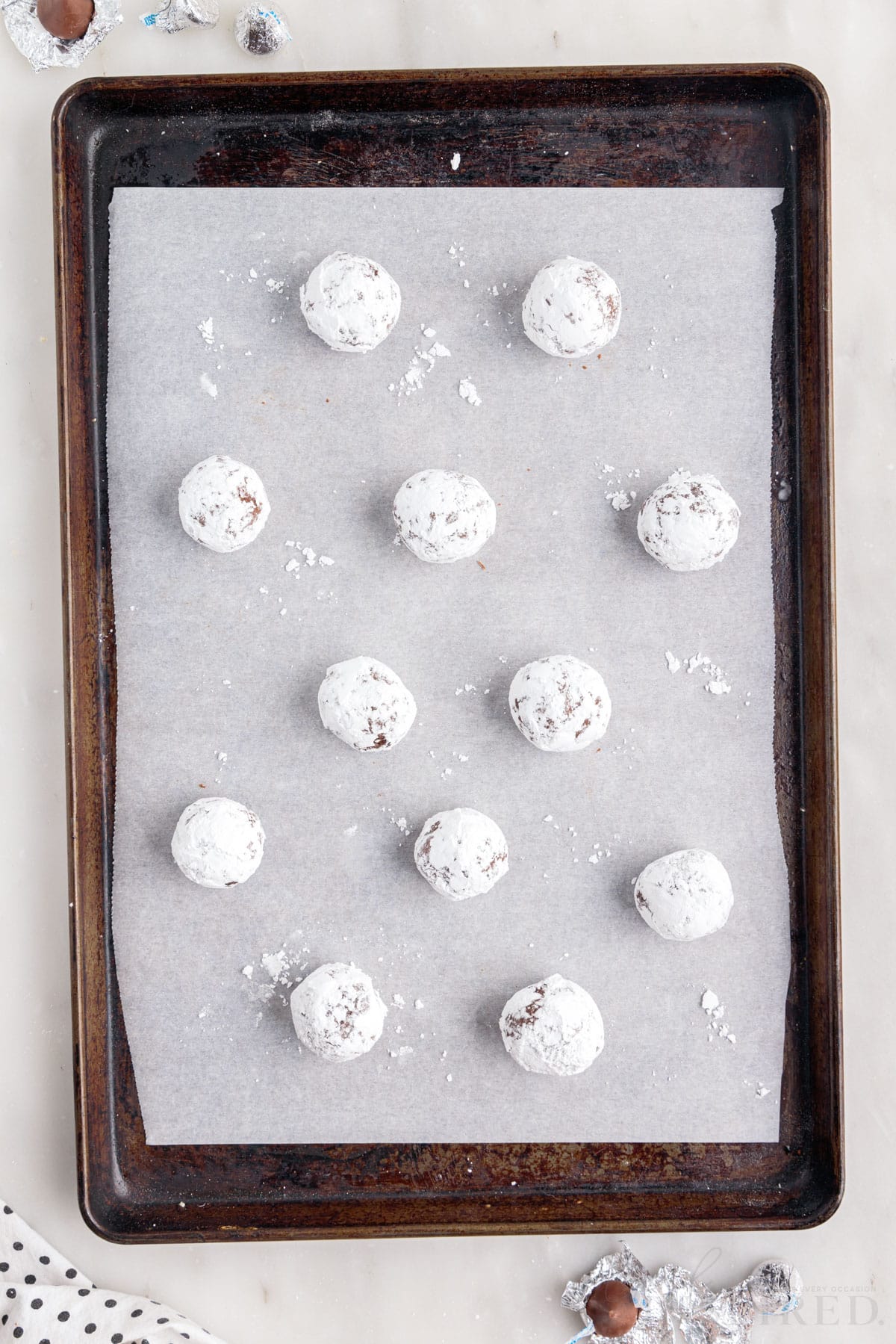 a cookie sheet lined with parchment paper with twelve chocolate crackle powdered sugar covered cookie dough balls