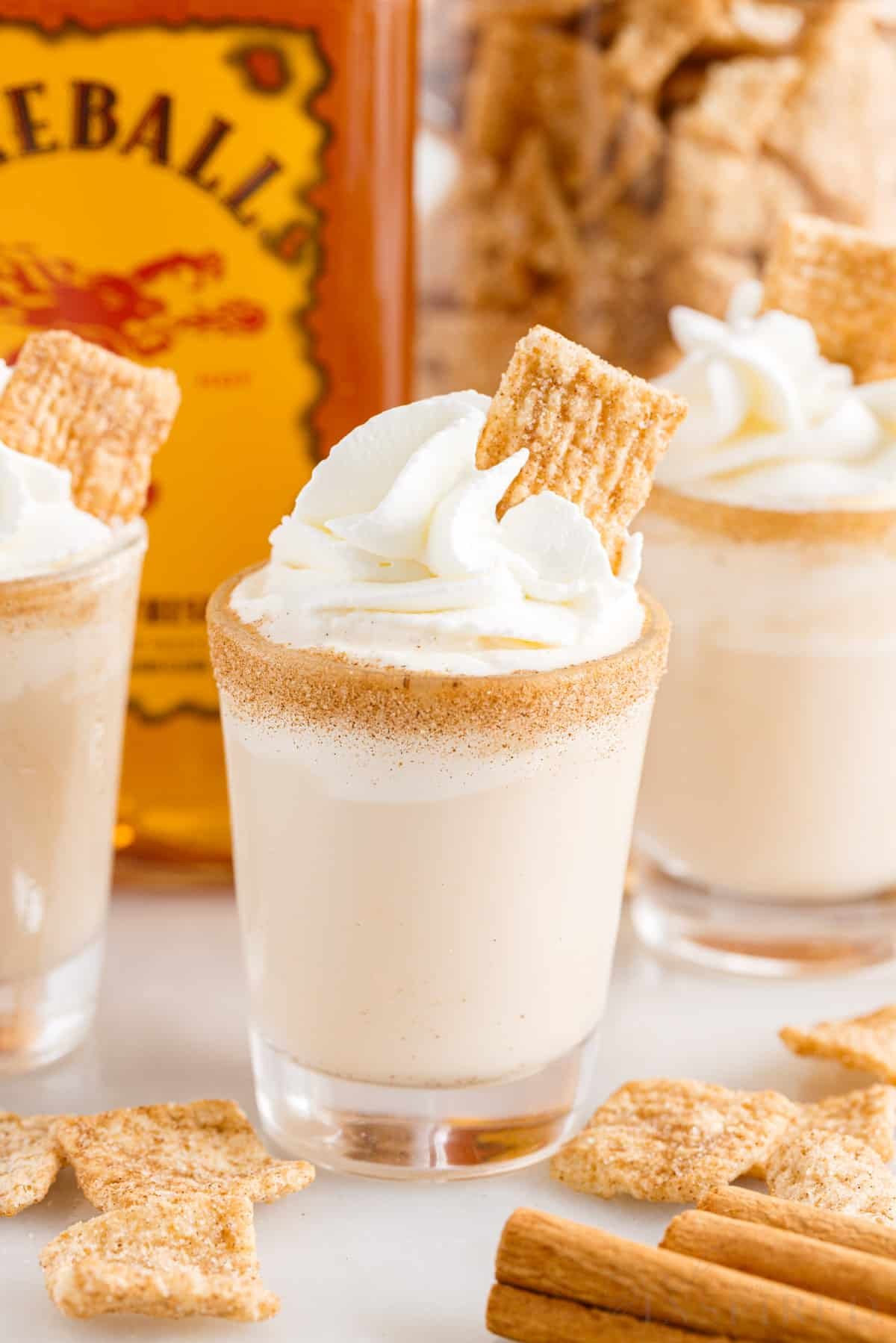 Cinnamon toast crunch shots topped with whipped cream and a piece of Cinnamon Toast Crunch cereal, Fireball whiskey in background.