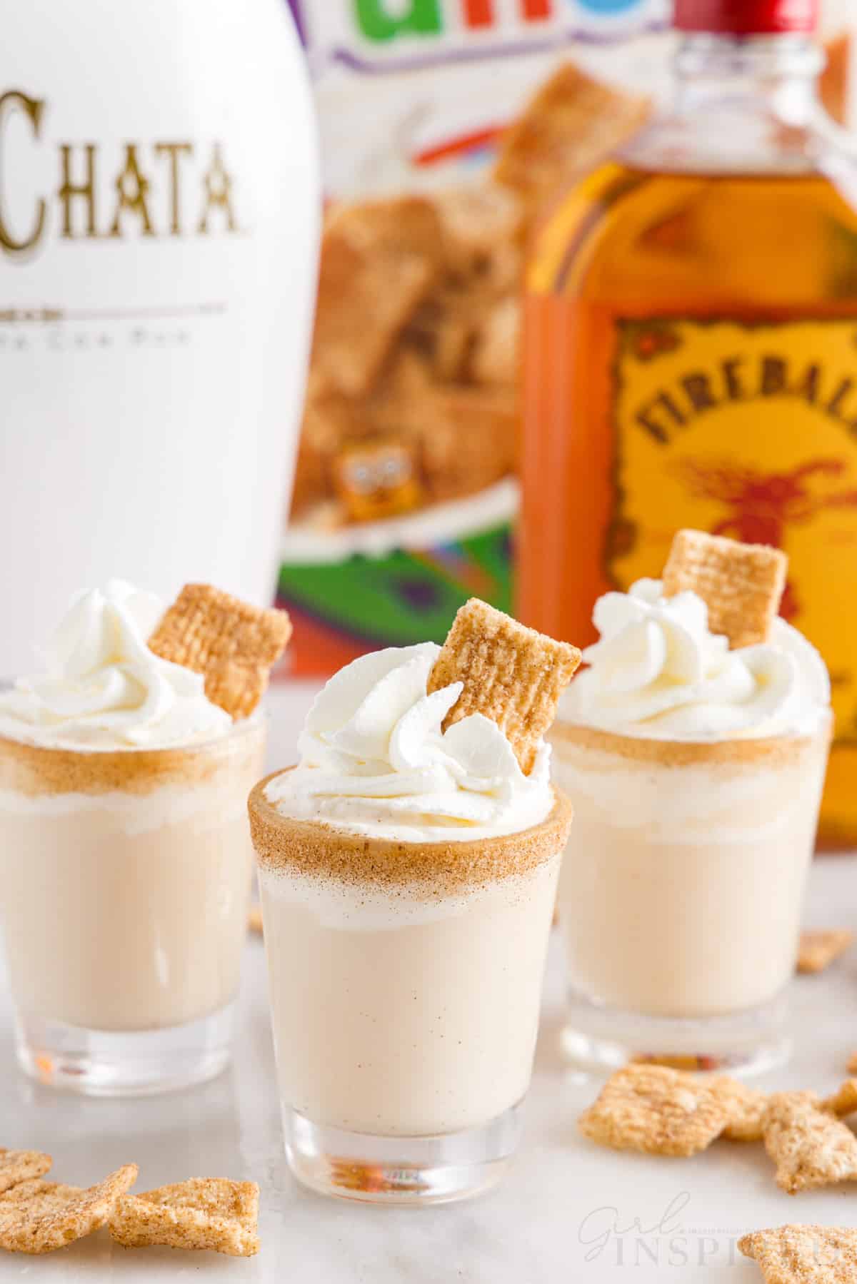 three finished cinnamon toast crunch shots with whipped cream and a piece of cinnamon toast crunch cereal in the top