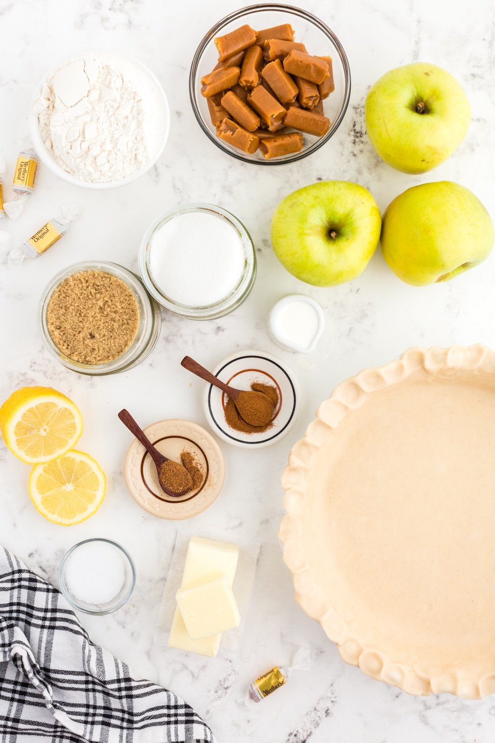 Pie crust pressed into a pie dish, black and white checkered linen, whole green apples, lemon halves, bowl of caramel candies, bowl of flour, bowl of light brown sugar, bowl of granulated sugar, small bowls of nutmeg and cinnamon, butter, on a white marble countertop.
