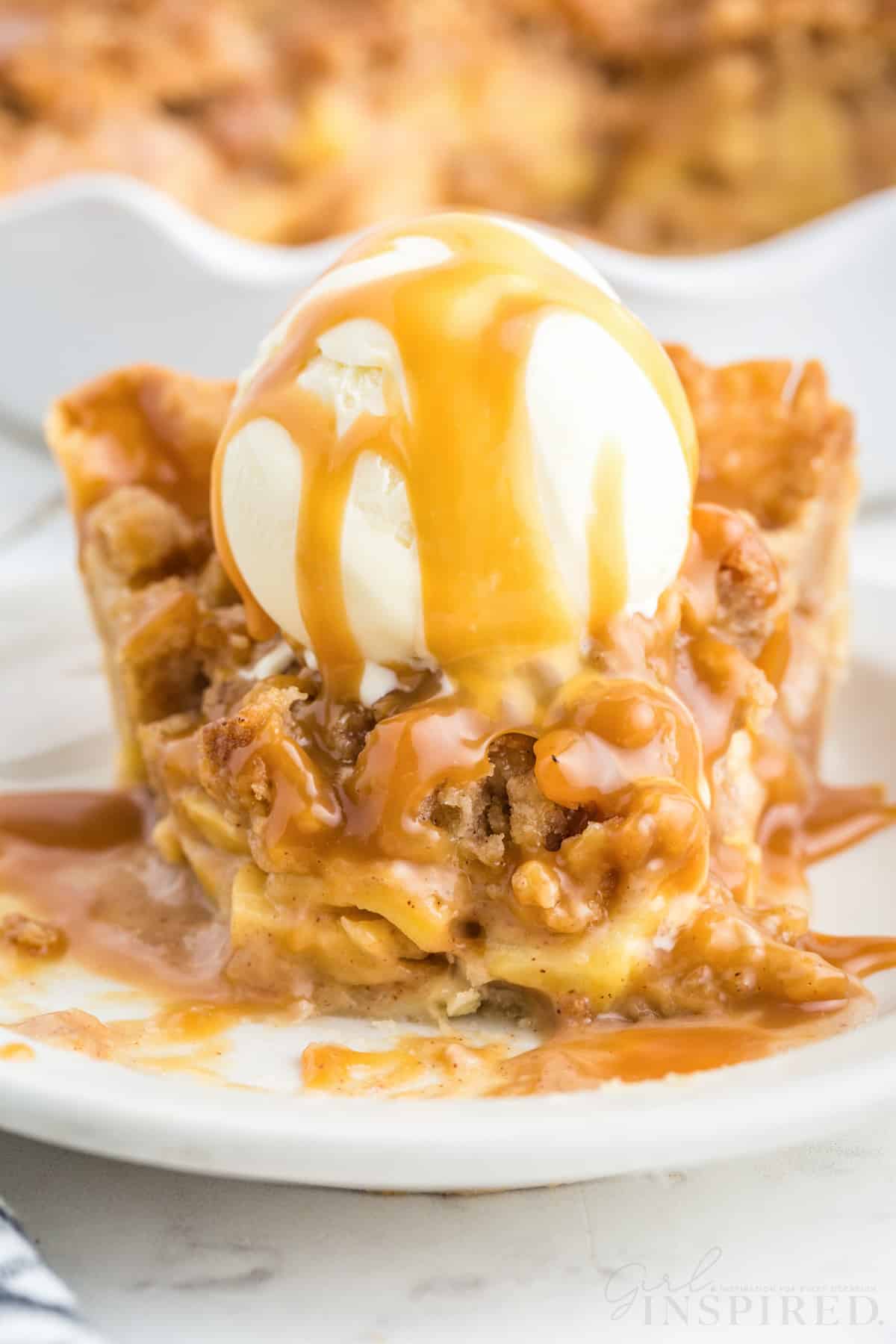 Slice of Dutch caramel apple pie on a white serving plate, scoop of vanilla ice cream on top of the apple pie slice, caramel apple pie in the background, checkered linen, on a white marble countertop.