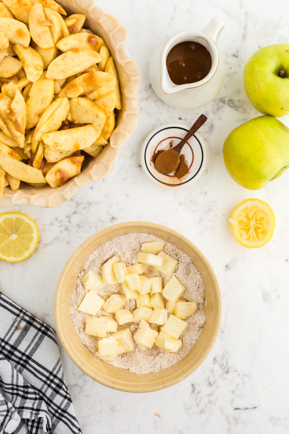 Bowl with dry streusel topping ingredients and cubed butter, black and white checkered linen, whole green apples, sliced lemons, jug of caramel sauce, small bowl of nutmeg and cinnamon, apple pie mixture in a prepared pie dish, on a white marble countertop.
