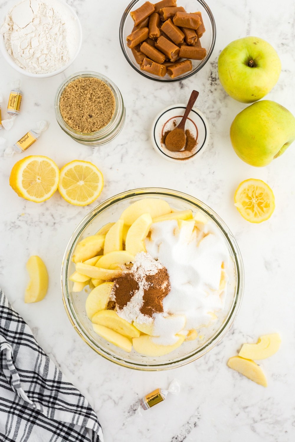 Large mixing bowl with sliced apples, flour, and spices, whole green apples, sliced lemons, black and white checkered linen, bowl of light brown sugar, bowl of granulated sugar, bowl of caramel candies, small bowl of nutmeg and cinnamon, on a white marble countertop.