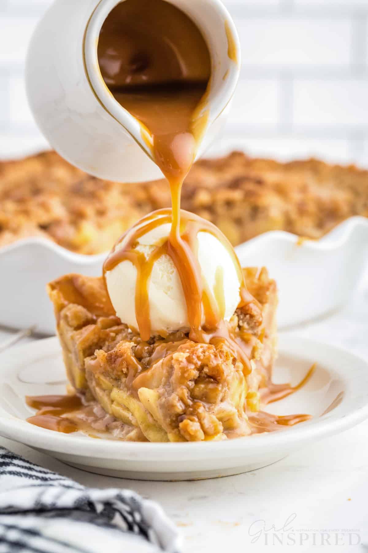 Caramel sauce drizzled over a slice of Dutch caramel apple pie on a white serving plate, Dutch caramel apple pie in a white pie dish, checkered linen, on a white marble countertop with white stacked tiles in the background.