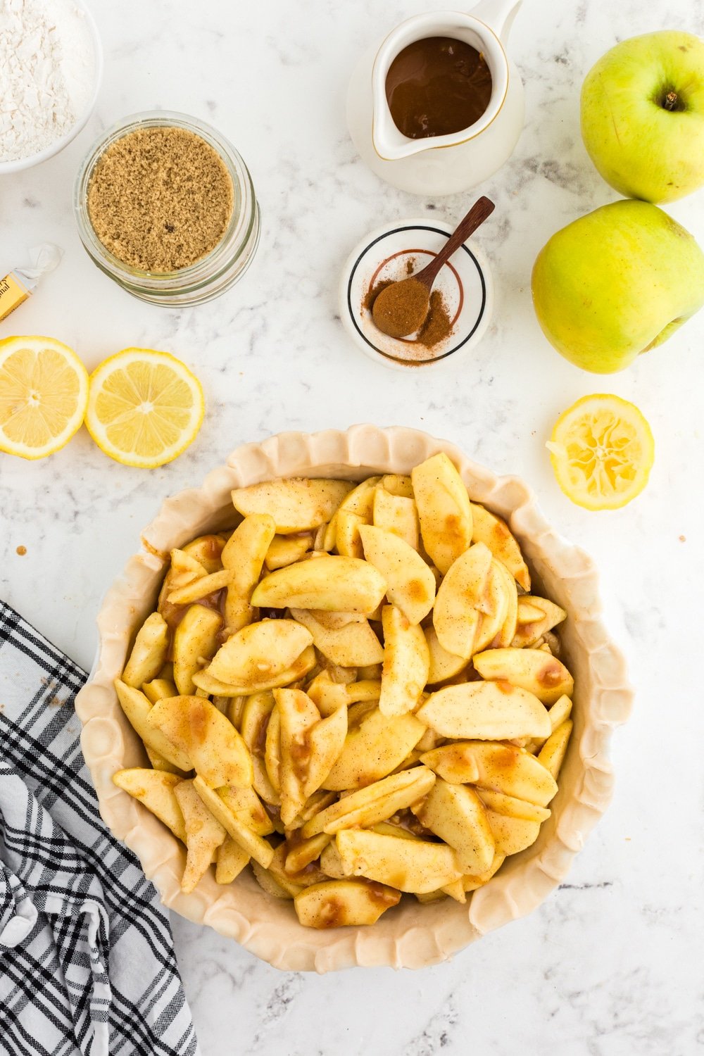 Apple pie mixture coated in caramel sauce added to a prepared pie dish with pie crust, black and white checkered linen, whole green apples, sliced lemons, jug or remaining caramel sauce, bowl of light brown sugar, bowl of granulated sugar, on a white marble countertop.