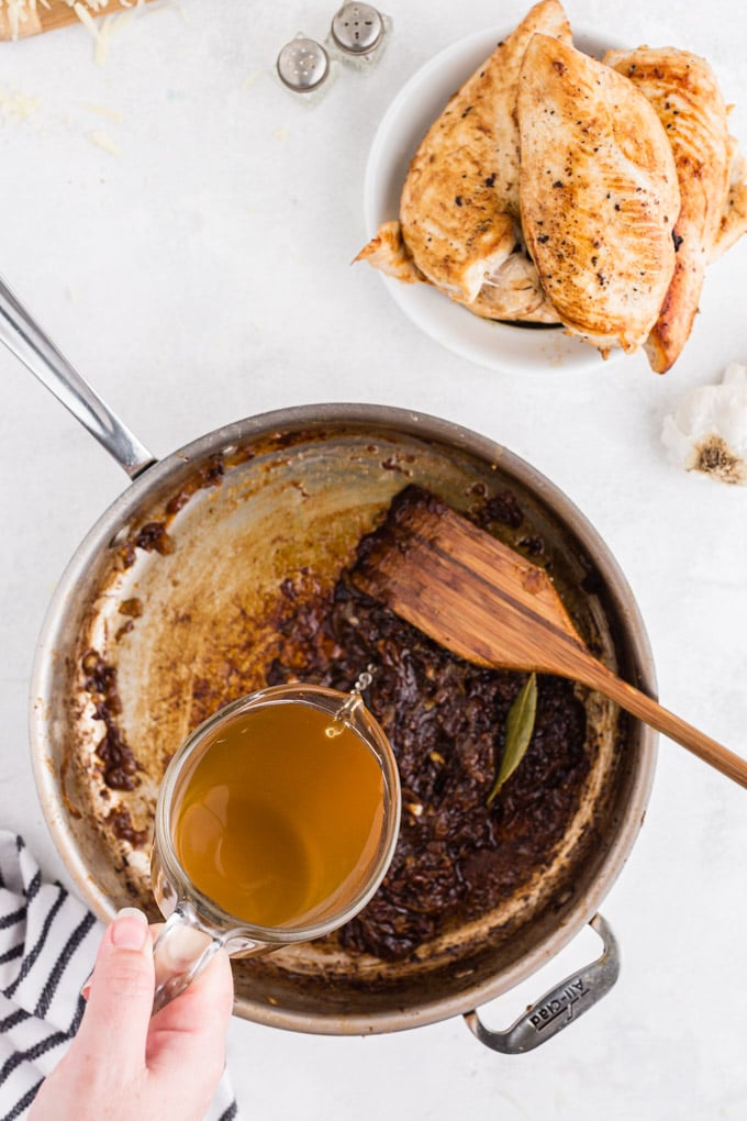 Onion gravy in a large skillet with beef broth being poured into it and a wooden stirring spoon, striped linen, plate of seared chicken breasts, on a white marble countertop.