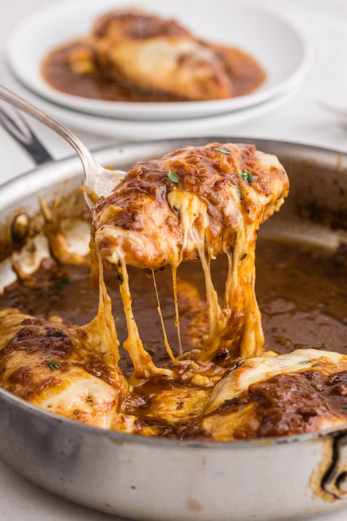 Metal serving spoon lifting a baked chicken breast with melted cheese and onion gravy from a large skillet, white serving plates in the background with another serving of French onion chicken, on a white marble countertop.